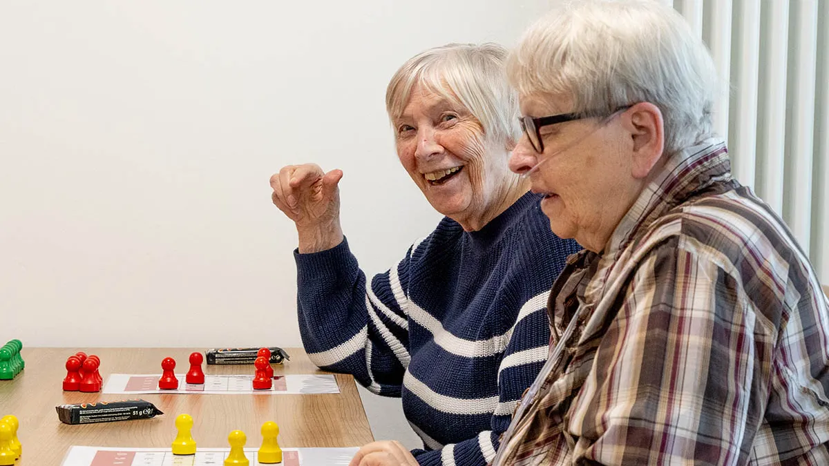 Zwei Seniorinnen sitzen an einem Tisch und spielen ein Gesellschaftsspiel bei PFLEGEN & WOHNEN UHLENHORST. Eine der Damen lacht in die Kamera.