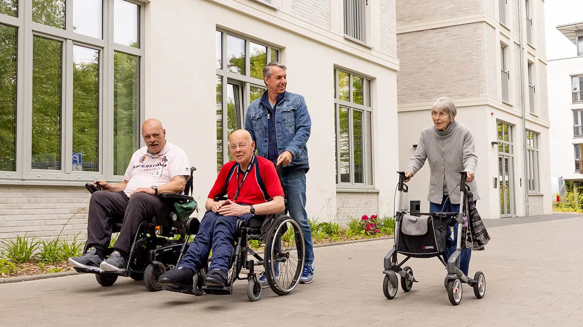 Eine Gruppe von zwei Rollstuhlfahrern, einer Dame mit Rollator und einem Mitarbeiter, der einen Rollstuhl schiebt, macht einen Spaziergang bei PFLEGEN & WOHNEN UHLENHORST.